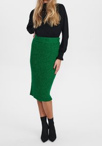 Grüne gerippte Strick-Midijupe mit figurbetontem Schnitt, kombiniert mit einem schwarzen Langarmoberteil. Schwarze struktierte Knöchelstiefel runden den Look ab.