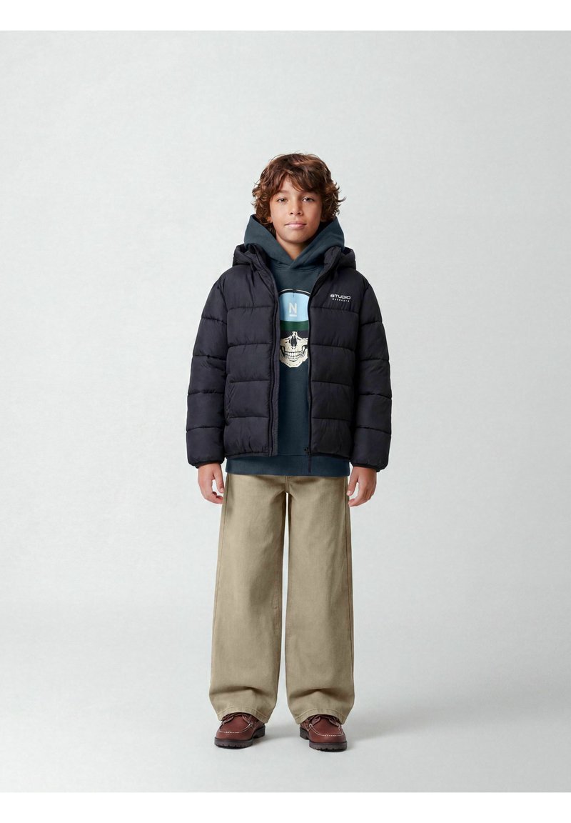 Garçon aux cheveux bouclés portant une doudoune noire, un sweat à capuche foncé avec un motif graphique, un pantalon large beige et des chaussures marron, debout devant un fond uni.