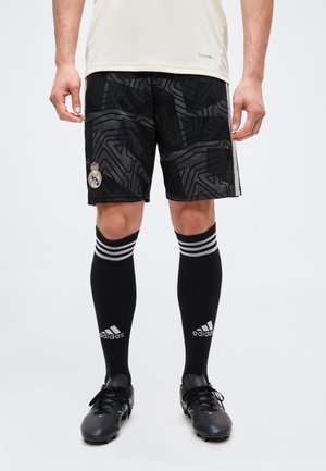 Mężczyzna noszący czarne spodenki piłkarskie Real Madrid z wzorem, czarne podkolanówki Adidas i czarne korki na białym tle.