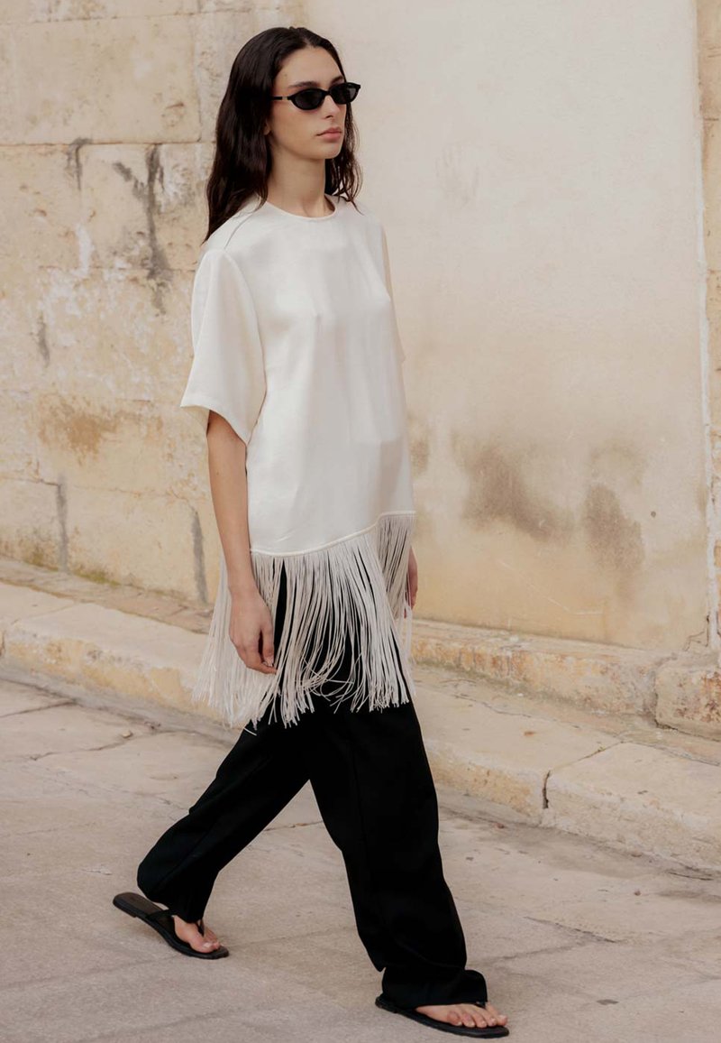Femme marchant à l'extérieur portant une chemise blanche à franges, un pantalon large noir, des tongs et des lunettes de soleil noires devant un mur en pierre beige.
