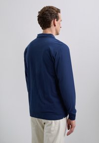 Hombre de perfil vistiendo una camisa de manga larga azul marino y pantalones beige frente a un fondo gris claro liso.