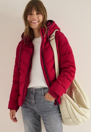 Doudoune rouge avec fermeture éclair sur le devant, capuche, design matelassé et poches latérales. Portée avec un pull blanc et un jean gris, assortie avec un sac crème.
