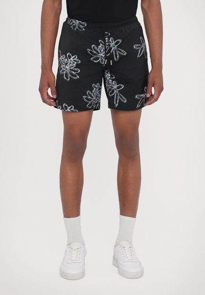 Shorts de bain noirs avec un motif floral argenté, taille élastique et cordon de serrage, fabriqués en tissu léger. Portés avec des baskets blanches et des chaussettes.