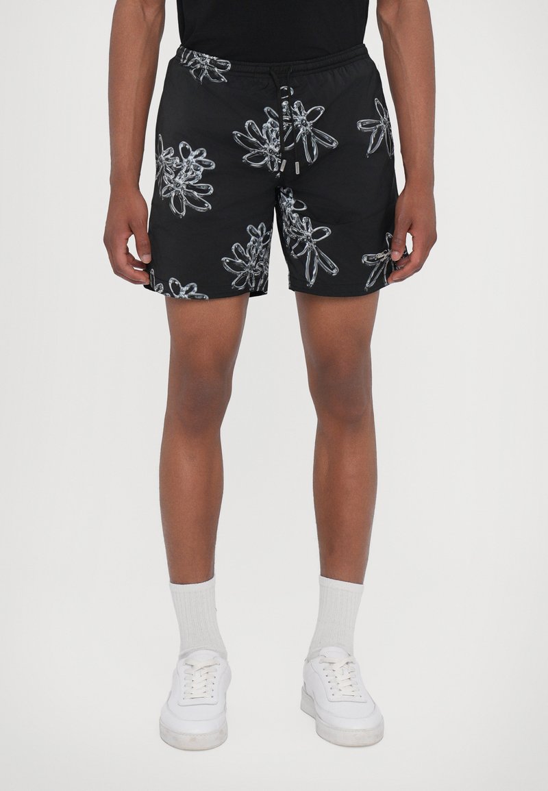 Schwarze Schwimmshorts mit einem silbernen Blumenmuster, elastischem Bund und Kordelzug, aus leichtem Stoff. Getragen mit weißen Sneakers und Socken.