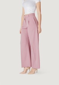 Pantaloni a vita alta e gamba larga in morbido tessuto rosa, caratterizzati da pieghe frontali e una texture liscia, abbinati a scarpe con tacco nude.