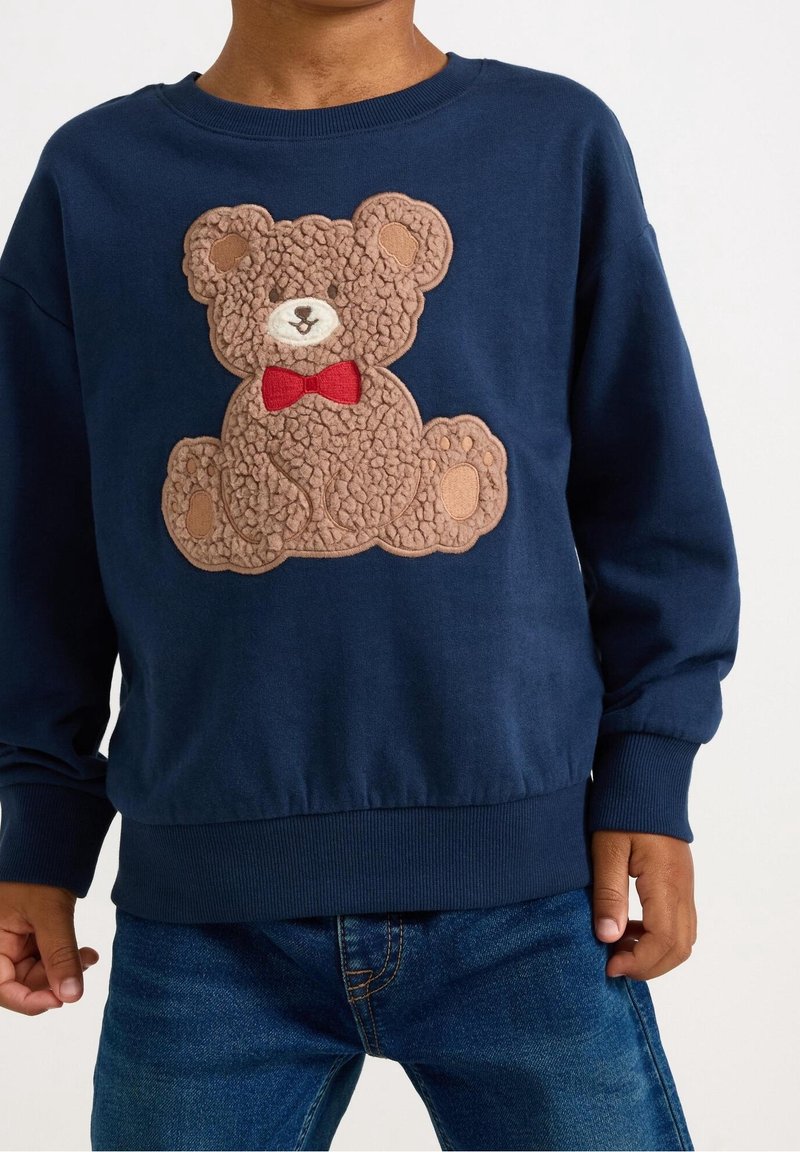 Enfant portant un sweat-shirt bleu marine avec un appliqué d'ours en peluche texturé et un nœud papillon rouge sur le devant.