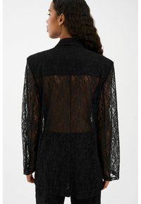 Melnas mežģīņu žakete ar caurspīdīgu aizmuguri, garām piedurknēm un pielāgotu piegriezumu. Vecums ar sarežģītiem ziedu rakstiem un iegriezta apkakle.