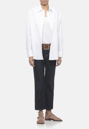 Persona che indossa una camicia bianca abbottonata sopra una canotta bianca, jeans neri a gamba larga, una cintura marrone e sandali marroni con borchie, in piedi su sfondo bianco.