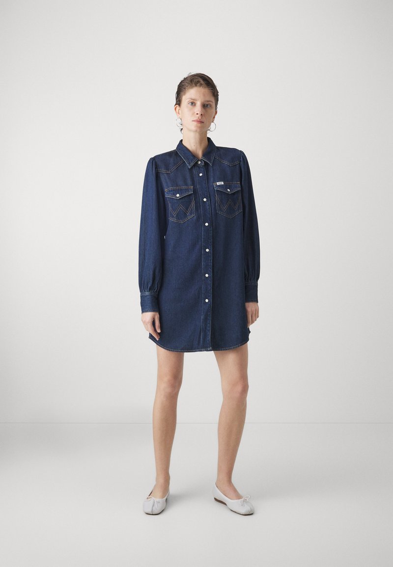 Robe chemise en denim bleu foncé avec de longues manches bouffantes, fermeture à boutons, deux poches poitrine et broderies décoratives sur les poches.