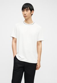 Νέος άνδρας με κοντά μαύρα μαλλιά στέκεται με το ένα χέρι στην τσέπη, φορώντας ένα λευκό μπλουζάκι με μαύρες λεπτομέρειες και μαύρο παντελόνι, μπροστά σε απλό φόντο.