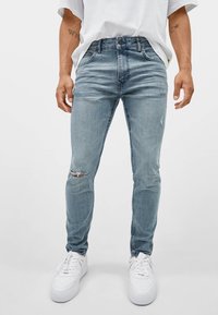 Jeans ajustados de color azul claro con detalles desgastados y un acabado descolorido. Hechos de mezclilla con una textura suave, combinados con zapatillas blancas.