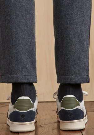Personne portant des chaussettes noires et un pantalon gris debout sur un sol en bois, des baskets blanches avec des accents verts et bleu marine à talon visibles.