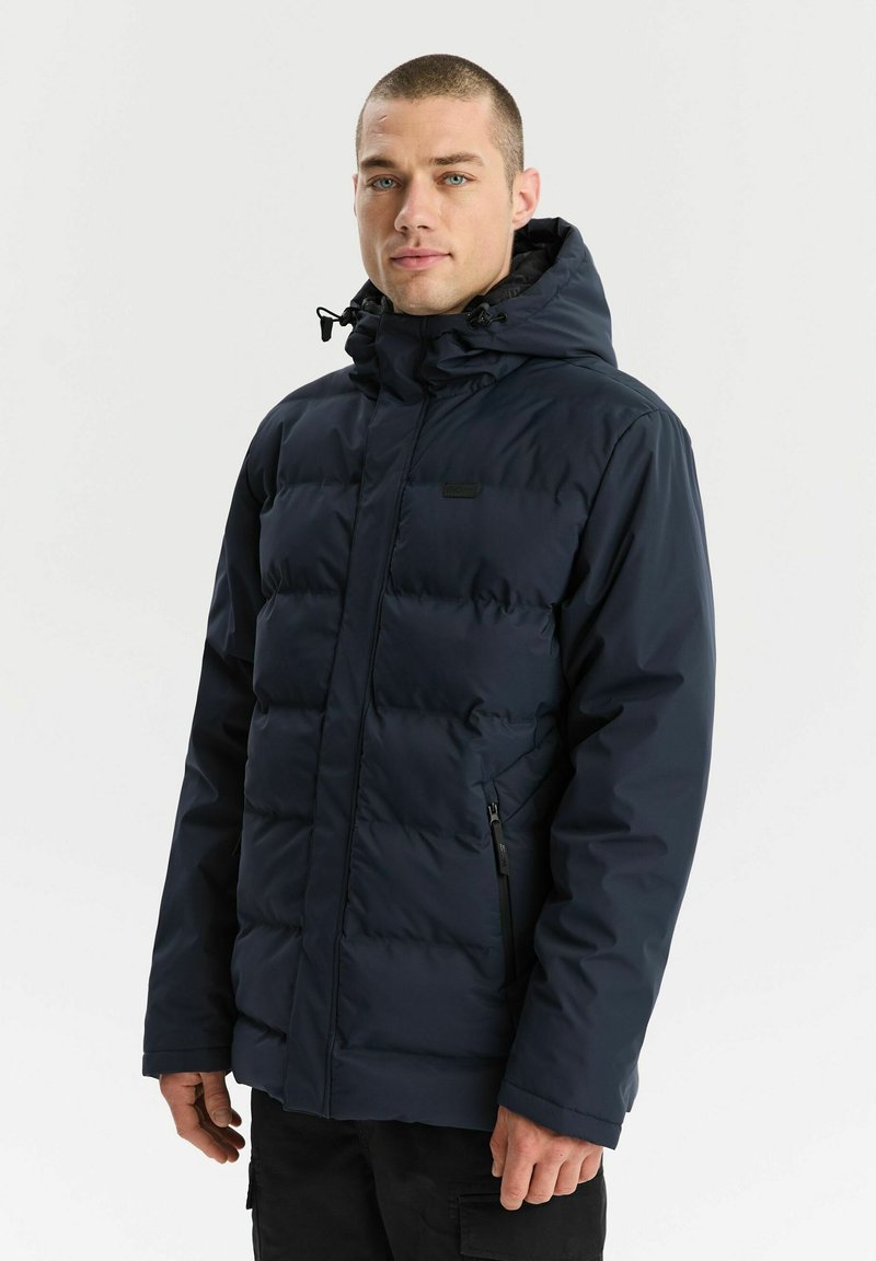 Mężczyzna z krótkimi włosami w granatowej kurtce puchowej z kapturem i czarnych spodniach, stojący na tle gładkiej białej ściany.