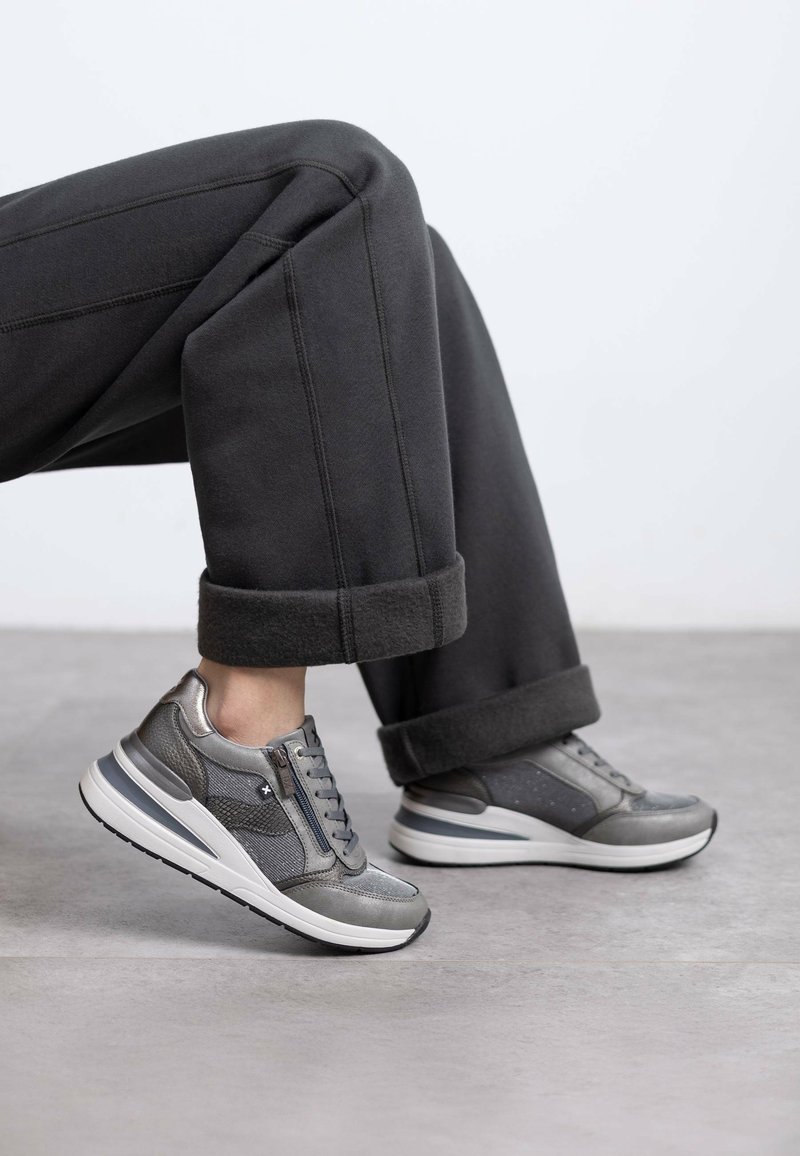 Zapatillas deportivas grises con una mezcla de materiales lisos y texturizados. Presentan un cordón en la parte frontal, acentos con cremallera y una suela gruesa blanca.