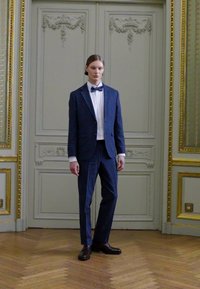 Κουστούμι με σκούρο μπλε σακάκι και ταιριαστά παντελόνια, συνδυασμένο με λευκό πουκάμισο και μαύρη παπιγιόν. Λείος υφή, επίσημος σχεδιασμός.