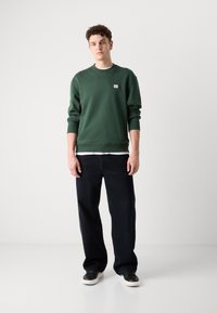 Grønn bomullsgenser med ribbestrikkede mansjetter og kant, brukt over en hvit skjorte. Kombinert med løse, mørke denimjeans og svarte sneakers.