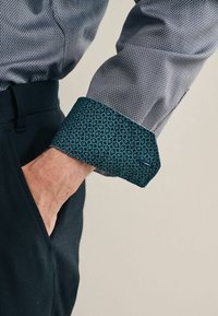 Skjorte med knapper og mønstret teal-ærme samt tekstureret grå stof. Ærmet har et blomstermotiv; skjorten har en klassisk pasform.