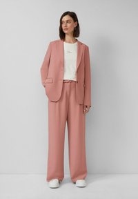 Vrouw in een stofroze pak met wijde broekspijpen en een blazer over een wit T-shirt, staande tegen een effen lichtgrijze muur.