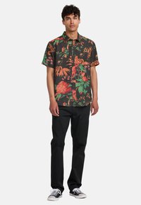 Kurzärmliges Blusenshirt mit einem dunklen Hintergrund und lebhaften roten und grünen Pflanzenmustern; kombiniert mit schwarzen, geraden Hosen.