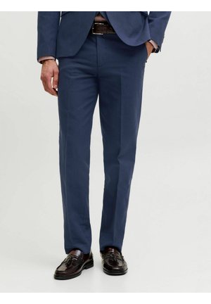 Férfi sötétkék öltönynadrágban, barna bőrszíjjal és sötétbarna bojtos mokaszinban, egyik kezét a zsebében tartva áll.