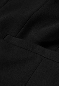 Gros plan sur un tissu noir avec une couture surpiquée et un détail de poche passepoilée.