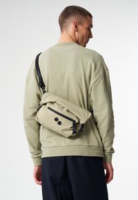 Beige Umhängetasche mit schwarzen Akzenten, Reißverschlusstasche, kreisförmigen Mustern und verstellbarem Tragegurt; aus strapazierfähigem, weichem Textil gefertigt.