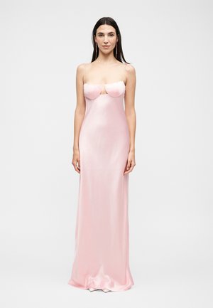 Γυναίκα που φοράει ένα αέρινο ροζ σατέν φόρεμα μήκους μέχρι το δάπεδο με στράπλες, δομητό μπούστο και λεπτές διάφανες τιράντες, στέκεται μπροστά σε λευκό φόντο.