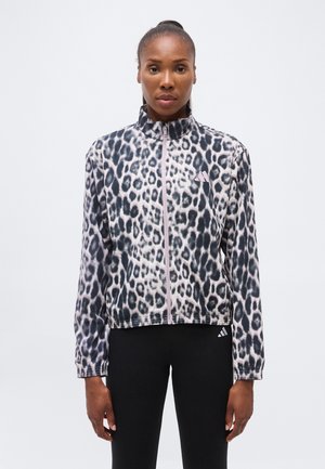 Femeie purtând o jachetă cu imprimeu leopard și fermoar, cu guler înalt și colanți negri, stând pe un fundal alb simplu.