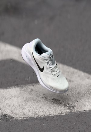 Chaussure de running Nike blanche avec logo noir flottant au-dessus d’un bitume gris aux marquages blancs.