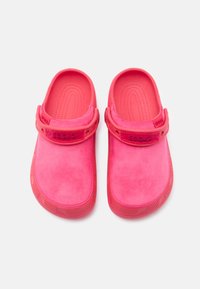 Une paire de Crocs roses à enfiler avec des dessus en texture suédine et des sangles de talon ajustables sur fond blanc.