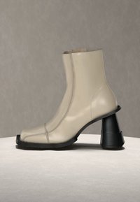 Μπεζ δερμάτινο μποτάκι αστραγάλου με ορατή ραφή μπροστά, τετράγωνη μύτη και φαρδιά, φουσκωτή μαύρη κυλινδρική φτέρνα σε ανοιχτή επιφάνεια.