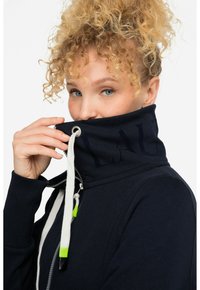 Sudadera con cremallera en color marino y cuello alto, con cordones en tonos verde y negro, y con el logotipo visible en el cuello.