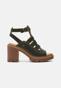 Sandale à talon en cuir vert foncé avec semelle épaisse marron, bout ouvert, multiples brides, boucle à la cheville et logo Timberland sur le côté.