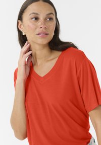 Červené tričko s výstřihem do V a krátkými rukávy, vyrobené z hladkého materiálu. Modelka se dotýká svého krku, má přirozený make-up a náušnice.