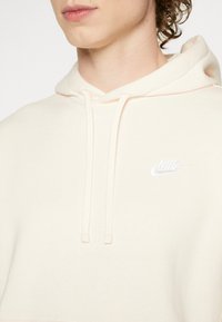 Krämfärgad Nike-hoodie med en ficka fram, dragsnoddar och en liten vit logo på vänster bröst. Mjuk, strukturerad tyg.