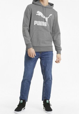 Haut à capuche en polaire gris avec une poche kangourou et un logo Puma blanc. Le modèle porte un jean bleu et des baskets noires avec des accents verts.