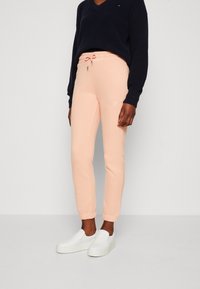 Persoon in lichtroze joggingbroek met elastische manchetten en een trekkoord, gecombineerd met witte instappers en een donkerblauwe trui.