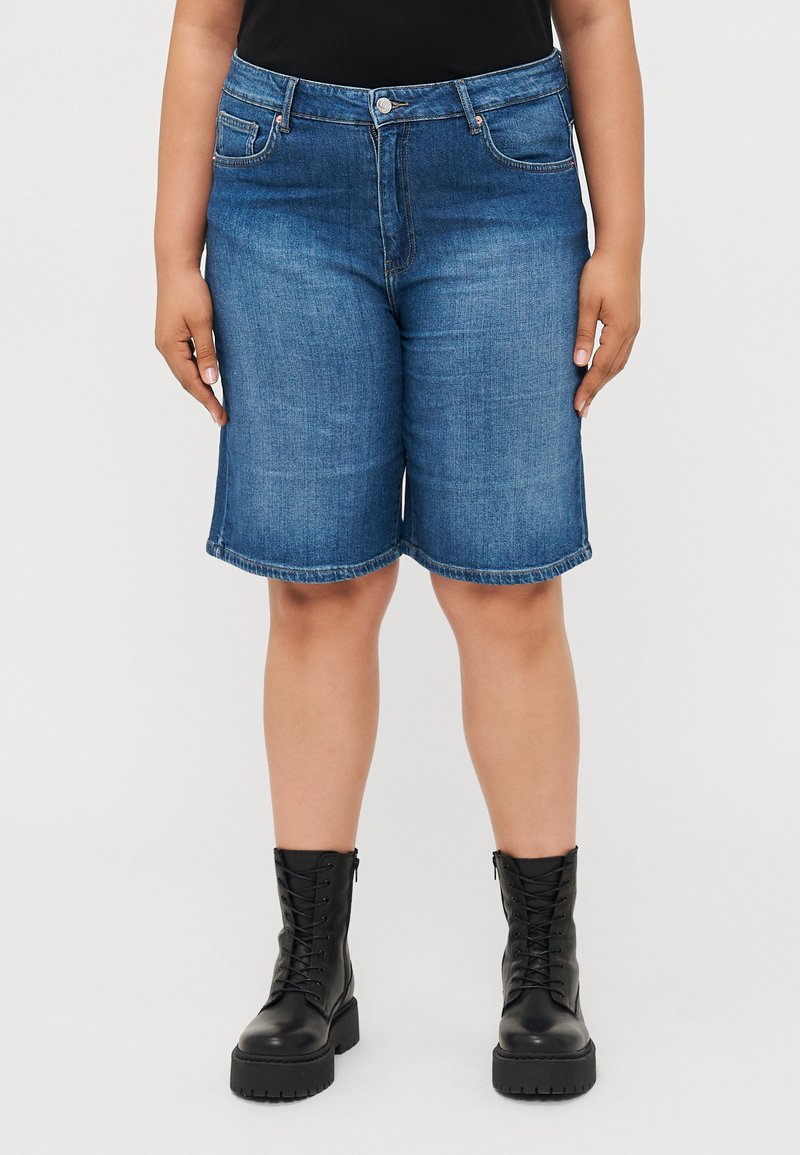 Sötétkék farmer Bermuda sort, egyenes fazonnal, öt zsebbel és felhajtott szegéllyel, fekete fűzős csizmával párosítva.