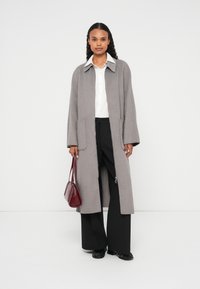 Pitkä harmaa villakangastakki, jossa on kaulus, sivutaskut ja rento istuvuus, yhdistettynä mustiin leveälahkeisiin housuihin ja viininpunaiseen käsilaukkuun.