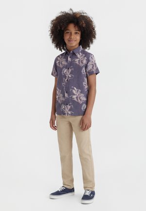 Chlapec s kučeravými vlasmi, oblečený v fialovej kvetinovej košeli s krátkym rukávom, béžových nohaviciach a tmavomodrých teniskách, stojí pred bielym pozadím.