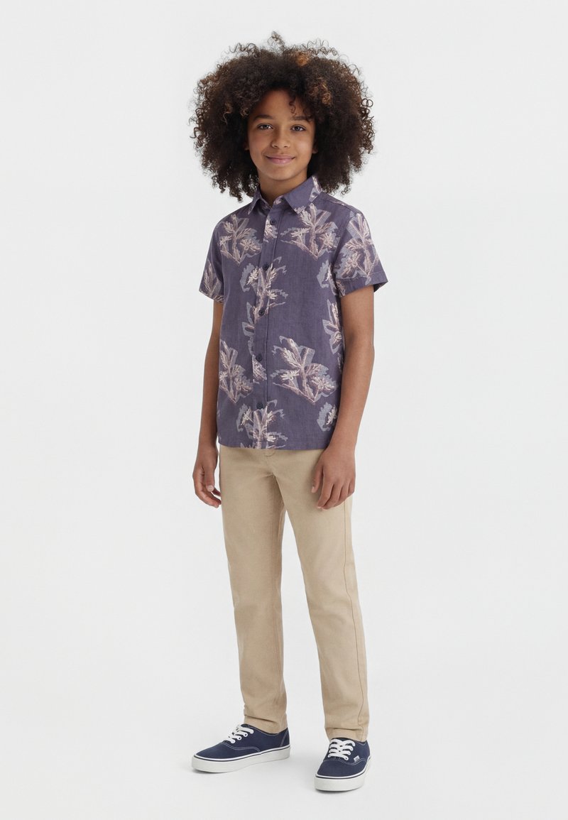 Αγόρι με σγουρά μαλλιά, ντυμένο με μωβ φλοράλ πουκάμισο κοντού μανικιού, μπεζ παντελόνι και μπλε σκούρα αθλητικά παπούτσια, στέκεται μπροστά σε λευκό φόντο.