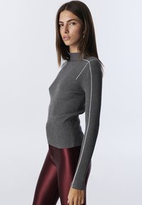 Woman with long brown hair wears a gray ribbed turtleneck sweater with white stripes and shiny maroon leggings, posing sideways.