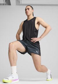 Sort ærmeløs atletisk tanktop og mørkegrå shorts, begge med Puma-logo. Hvide sneakers med pink og gule accenter.