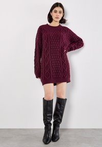 Burgunderfarbendes Strickkleid mit strukturiertem Rautenmuster, gerippten Bündchen und Saum; kombiniert mit knielangen schwarzen Lederstiefeln.