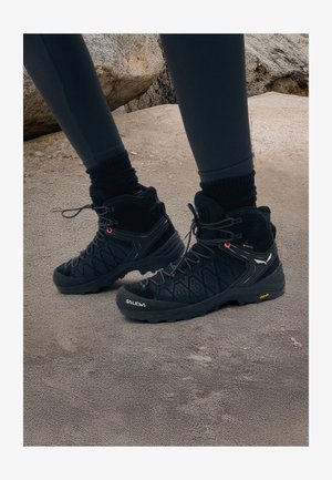 Bottes de randonnée noires avec une conception à cheville haute, surface texturée, bout renforcé et semelle Vibram. Présente un système de laçage et un marquage sur le côté.