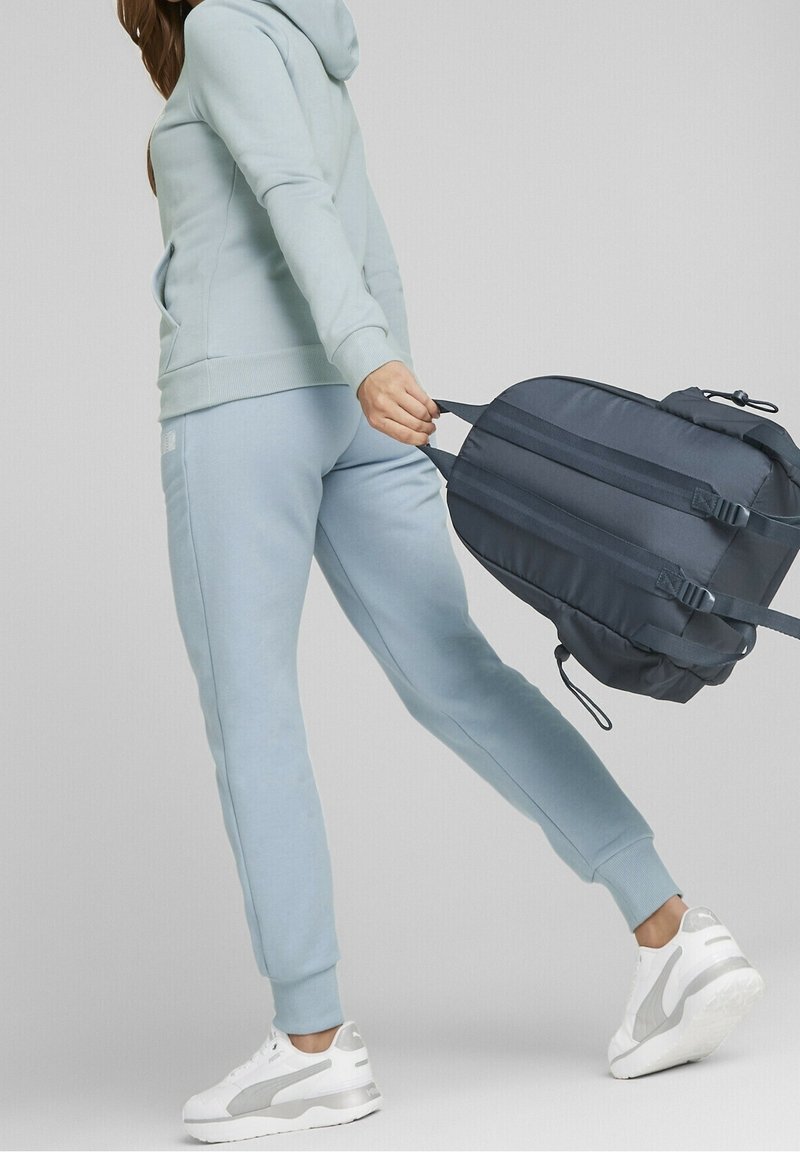 Sudadera azul claro y joggers a juego, de mezcla de algodón, de corte relajado, con una mochila azul oscura de dos asas, llevada en una mano.