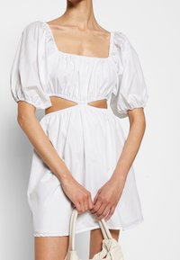 Vit puffärmad klänning med fyrkantig halsringning och utskärning vid midjan, tillverkad av ett mjukt, lätt tyg. Modell som håller en väska.