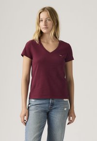 Ξανθιά γυναίκα με μαρουν κόκκινη μπλούζα V-neck και ανοιχτό μπλε τζιν με φθορές, στέκεται μπροστά σε απλό φωτεινό φόντο.