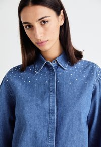 Denim shirt with a collared neckline, featuring a light blue color and scattered clear rhinestone accents on the shoulders.