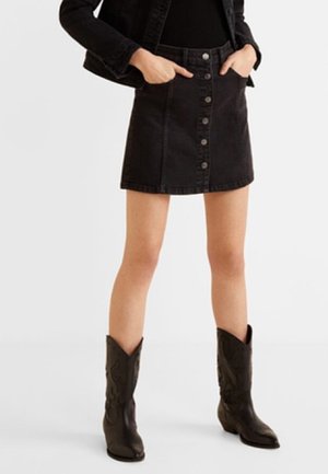 Personne portant une jupe en denim noire boutonnée à l'avant, un haut noir, une veste noire et des bottes de cowboy noires, debout les mains dans les poches de la jupe.